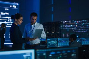Project Leader and Chief Executive Discuss Data Shown on a Laptop. In Background Big Screen Showing Infographics of Infrastructure, Charts, System Analysis. Telecommunications Control Room (Project Leader and Chief Executive Discuss Data Shown on a La